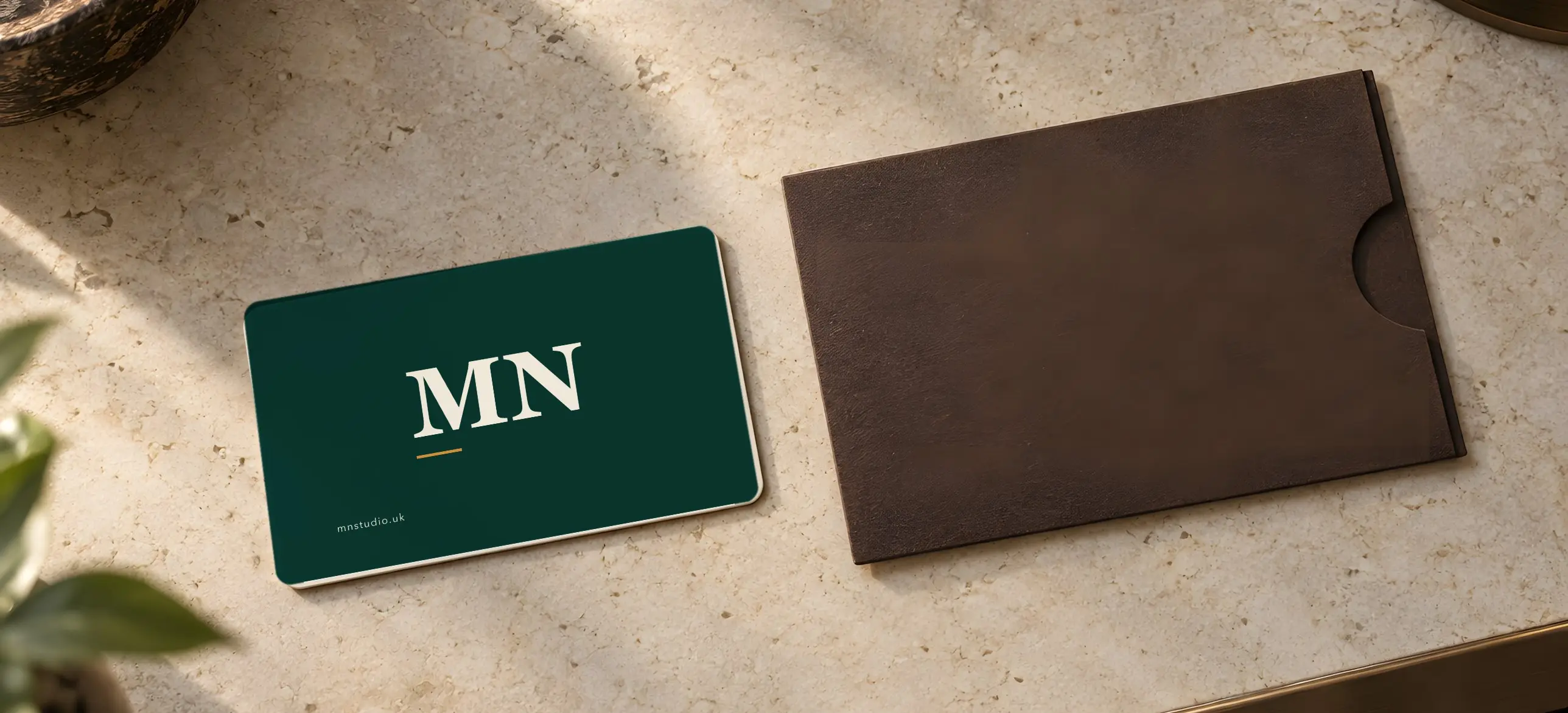A green MN Studio business card with the MN monogram beside a brown leather card sleeve on a stone table
