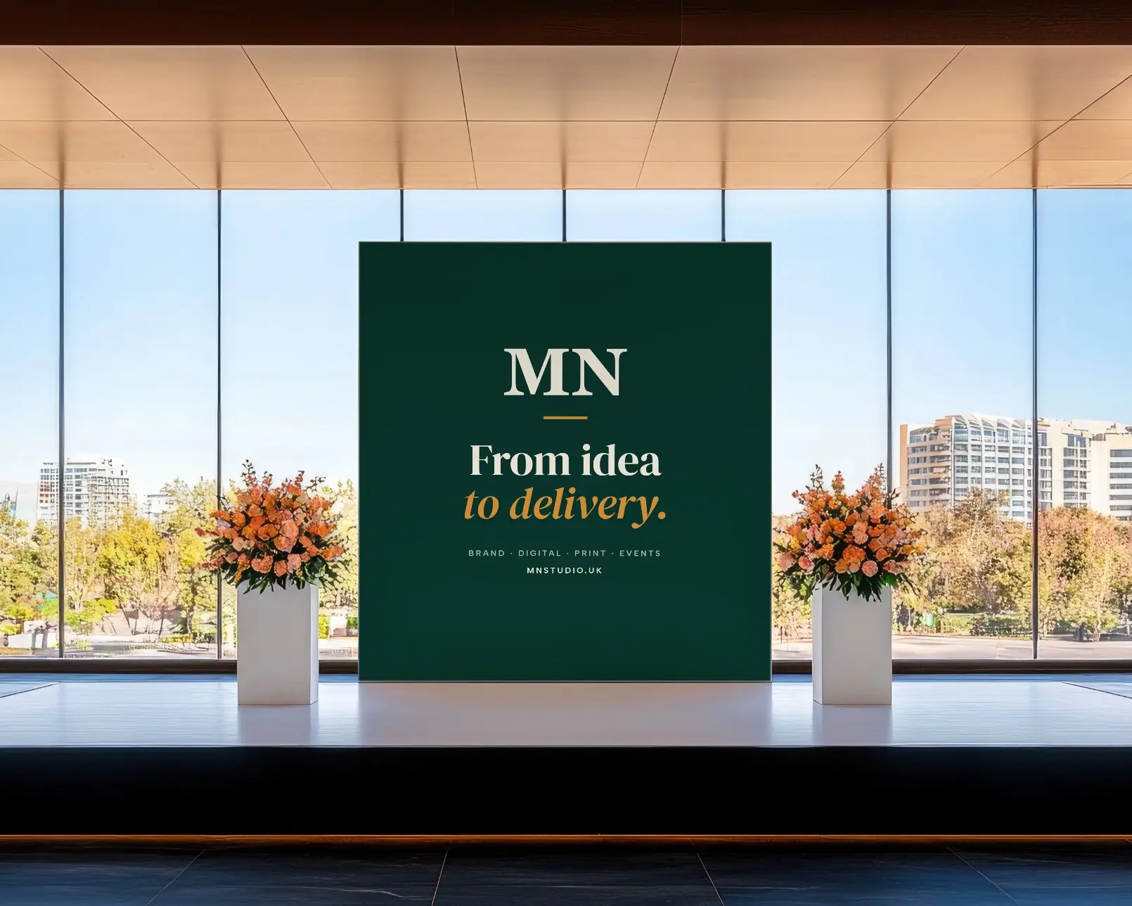 A stage in a bright glass-walled venue with a green MN Studio backdrop reading “From idea to delivery.” between two floral arrangements