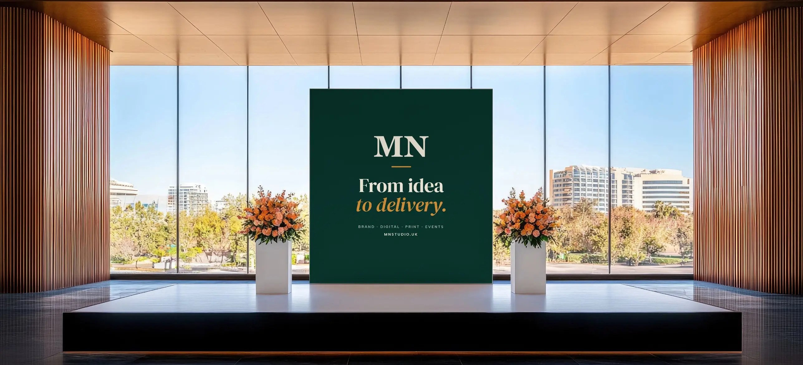 A stage in a bright glass-walled venue with a green MN Studio backdrop reading “From idea to delivery.” between two floral arrangements